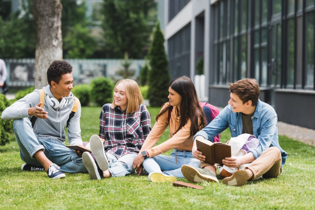 Group of teenagers wearing backpacks do their homework while sitting on the lawn outside of school