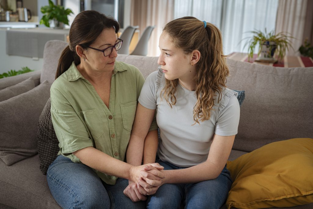 Comforting mother holds her teen daughter's hand as they sit on the living room sofa