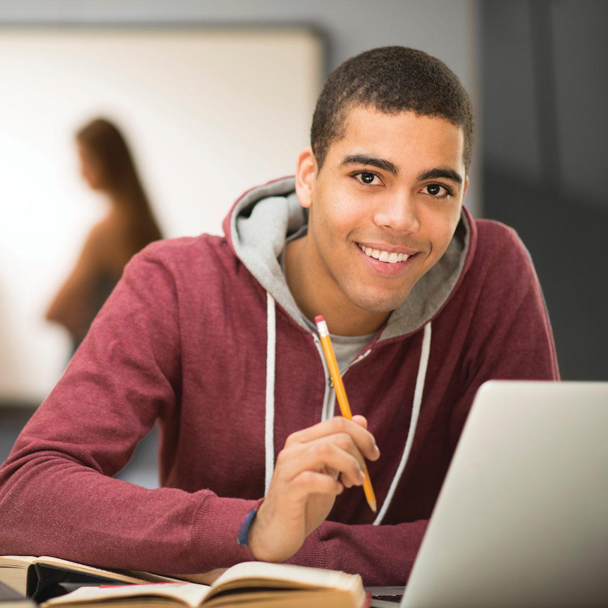 A teenage boy smiles as he holds a pencil and uses his laptop for school