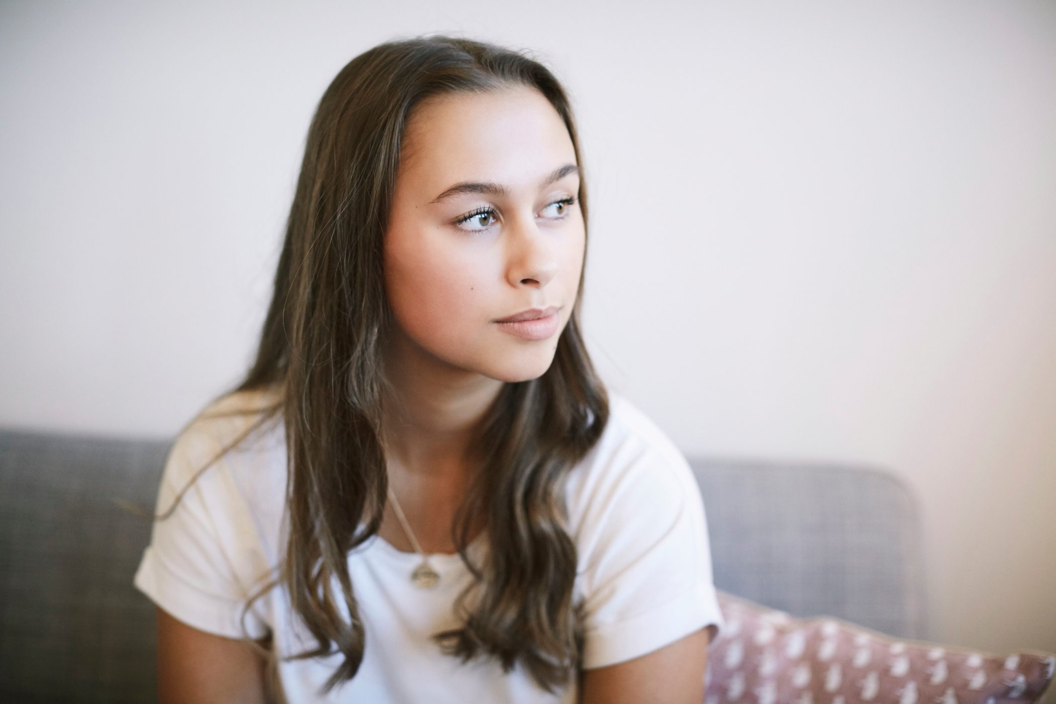 Teen girl gazes to her left while sitting on a grey sofa.