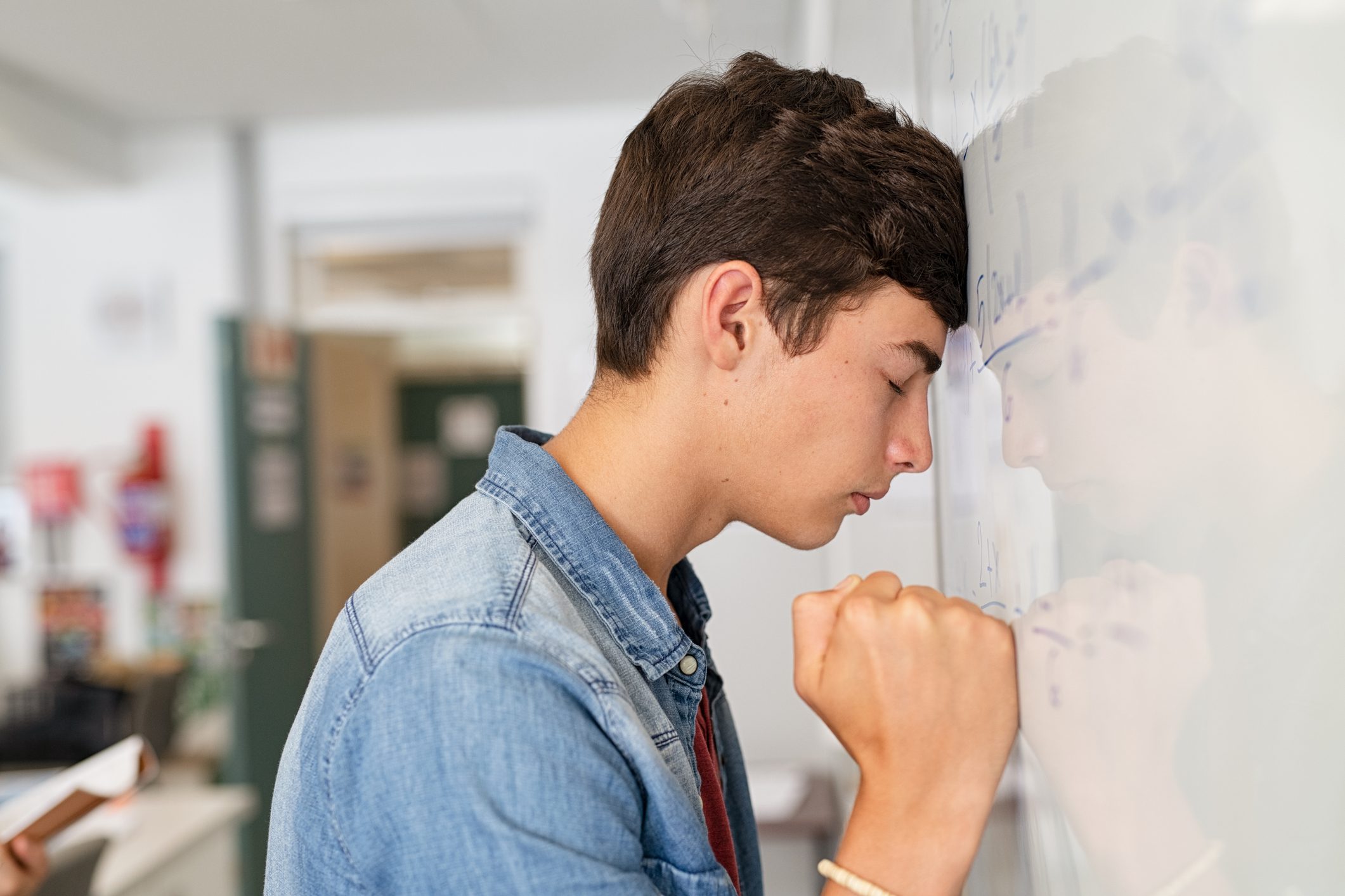 Stressed high school boy struggles with math assignment leans his head on whiteboard.